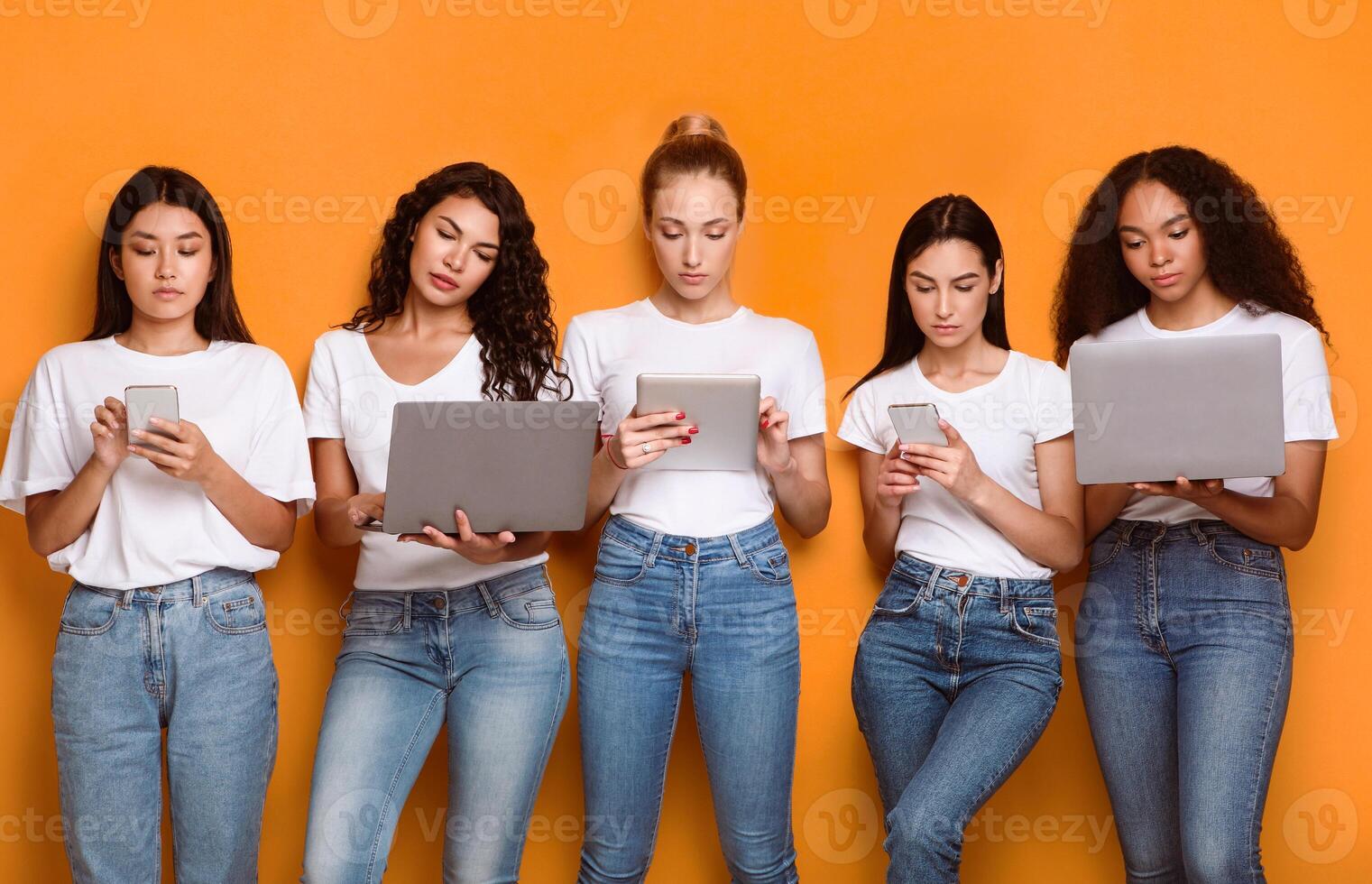 Five Diverse Ladies Using Modern Gadgets, Phones, Laptop And Digital Tablet Computers Browsing Internet Standing On Yellow Studio Background. Females Group Networking In Social Media Online photo