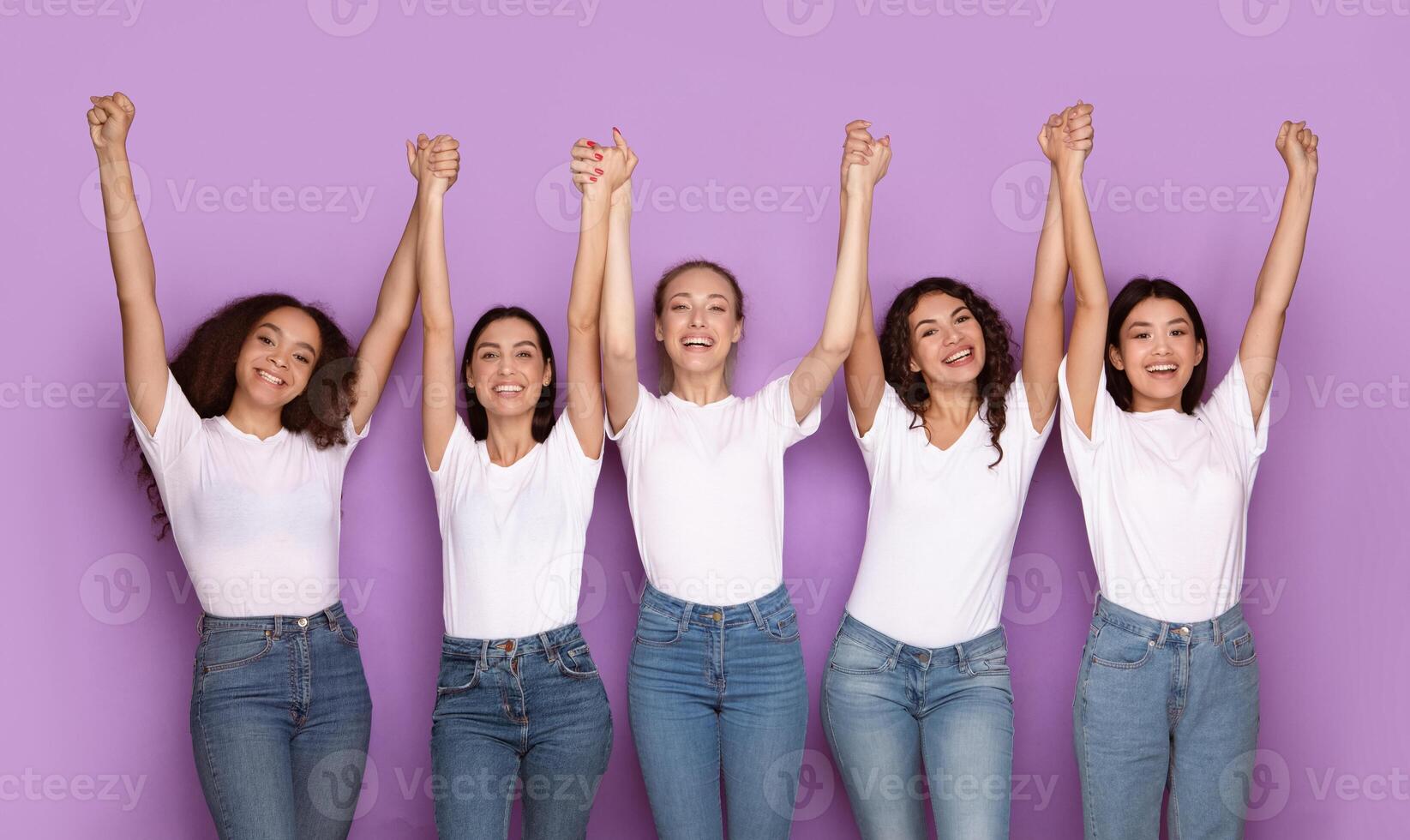Women's Power And Unity. Cheerful Multiethnic Ladies Raising And Holding Hands Posing Together Over Purple Studio Background, Smiling To Camera. Females Togetherness Concept photo