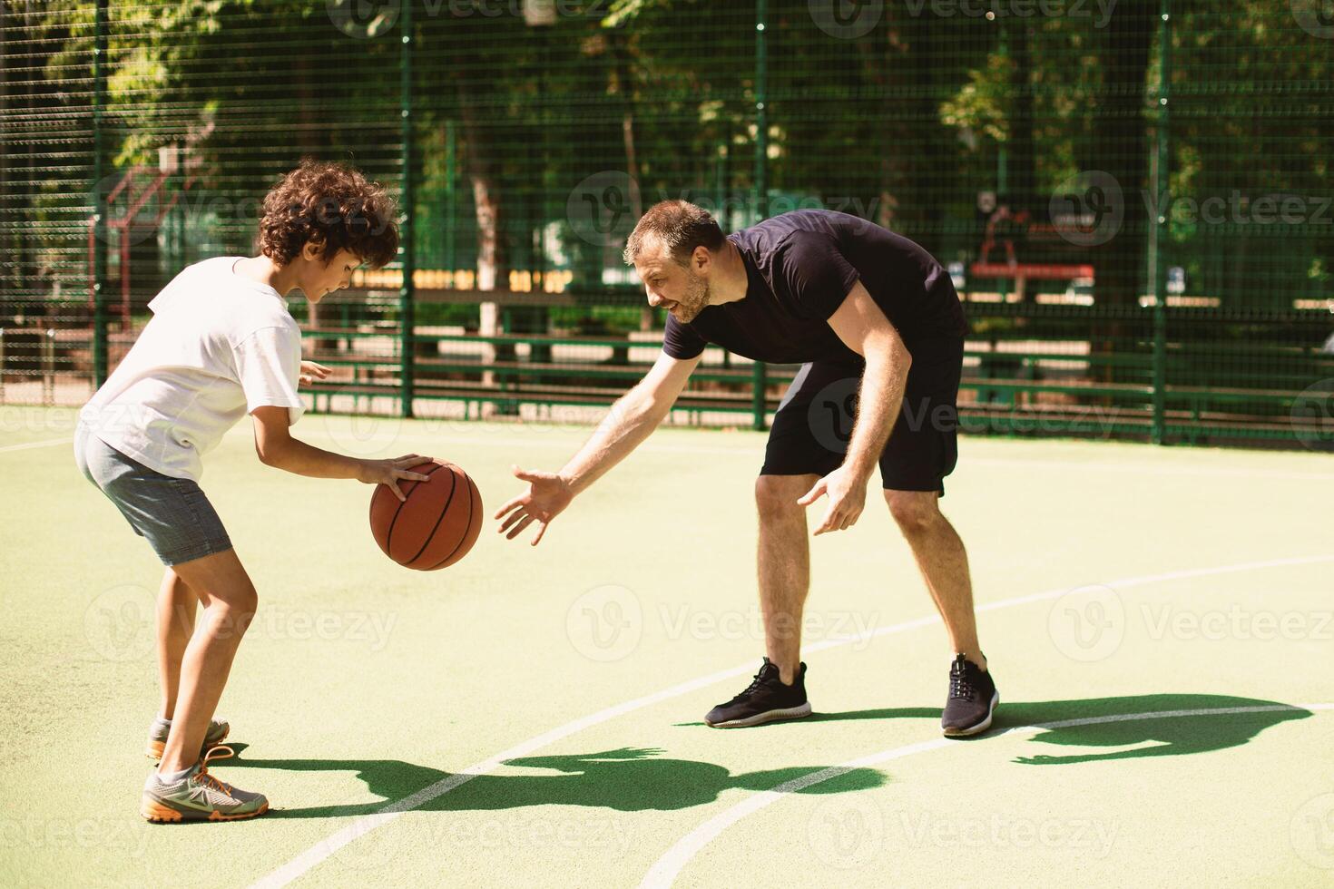 Children Wellness And Wellbeing. Portrait in action of sporty adult man and young focused boy playing basketball on a court with green grass outdoors in the summer on a sunny day. Hobby Concept photo