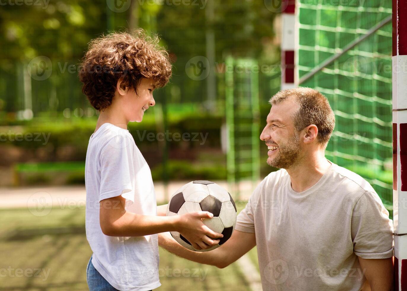 Active Lifestyle Concept. Curly boy holding soccer ball, playing with his dad on stadium, side view photo