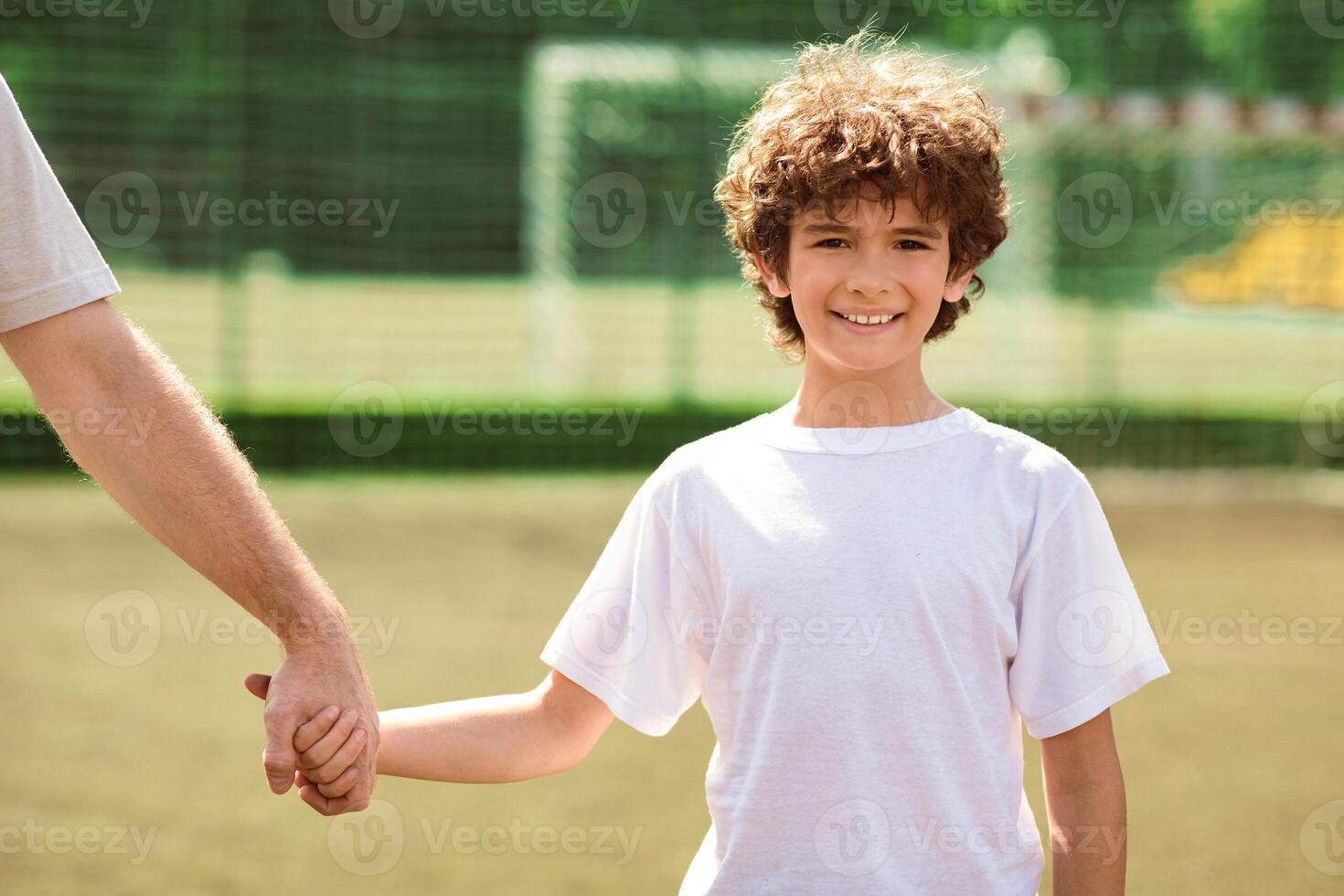 Support Concept. Portrait of smiling curly boy holding male hand and looking at camera, standing on football field photo