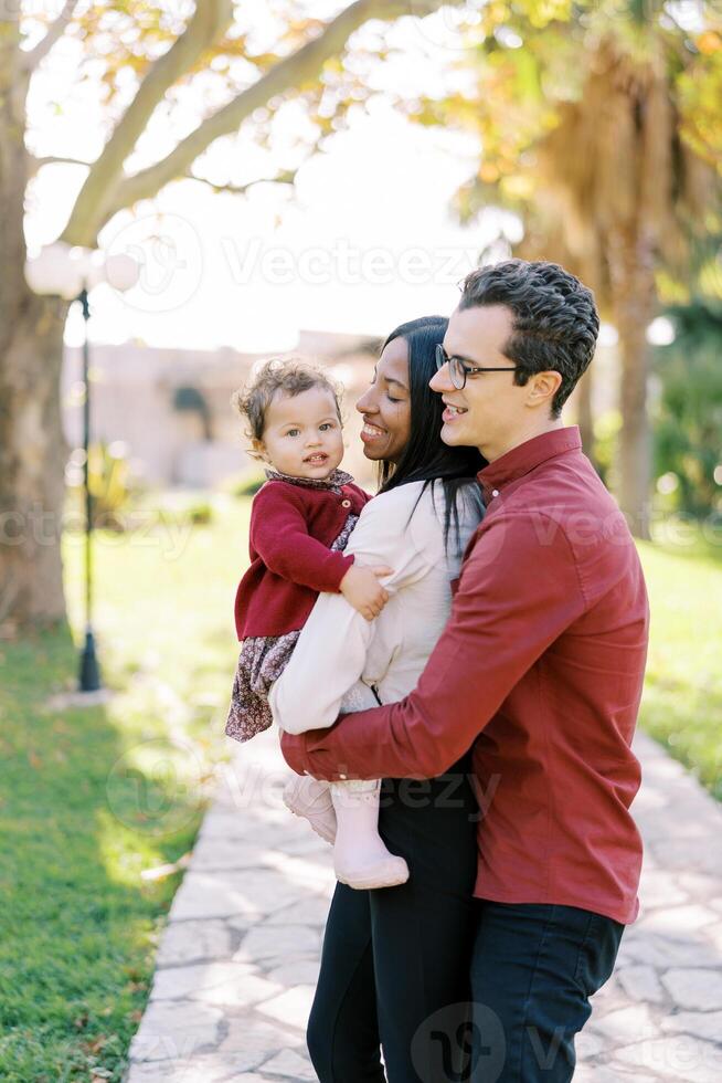 Dad hugging mom from behind with a little girl in her arms in the park photo