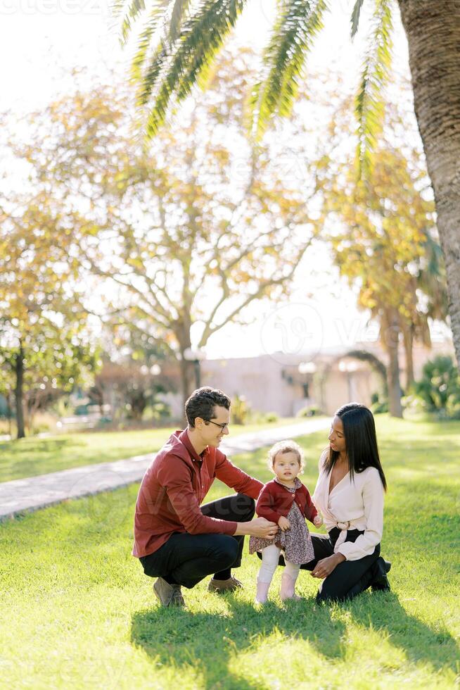 Smiling parents squatting and looking at little girl in sunny park photo