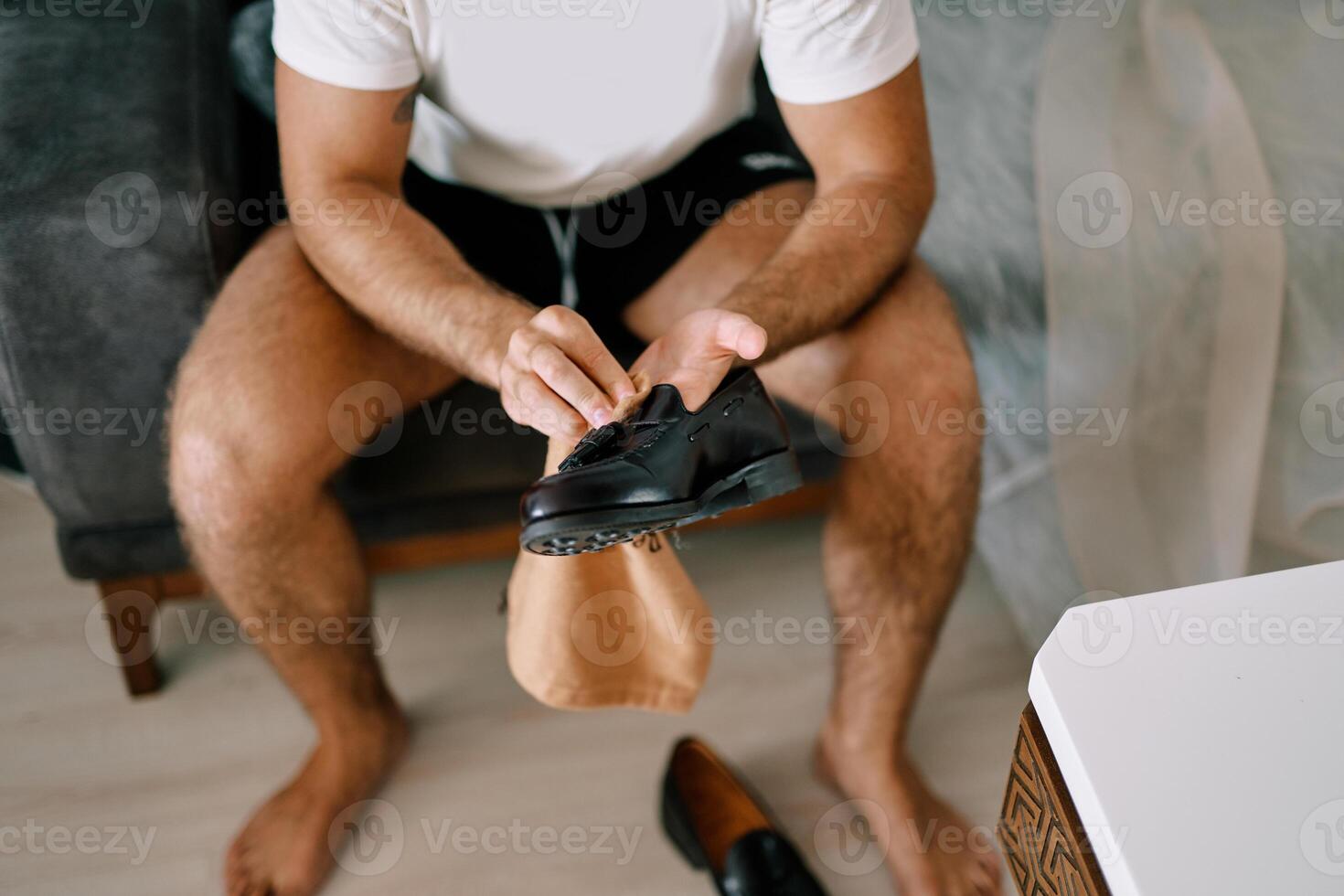 Barefoot man sits on the bed and wipes his black leather shoes with a napkin. Cropped. Faceless photo