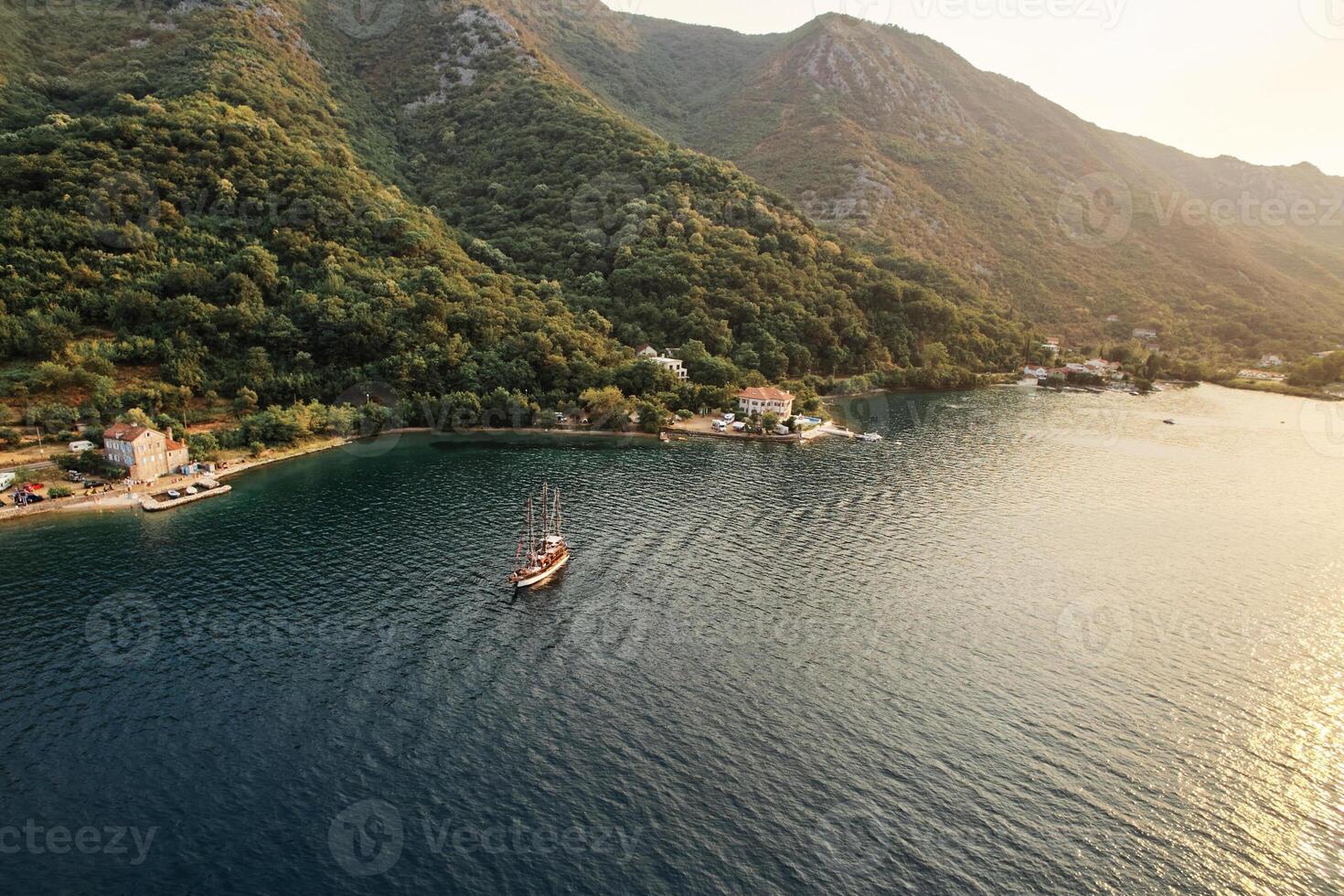 Sailboat sails across the bay against the backdrop of a green, wooded mountain coast. Drone photo