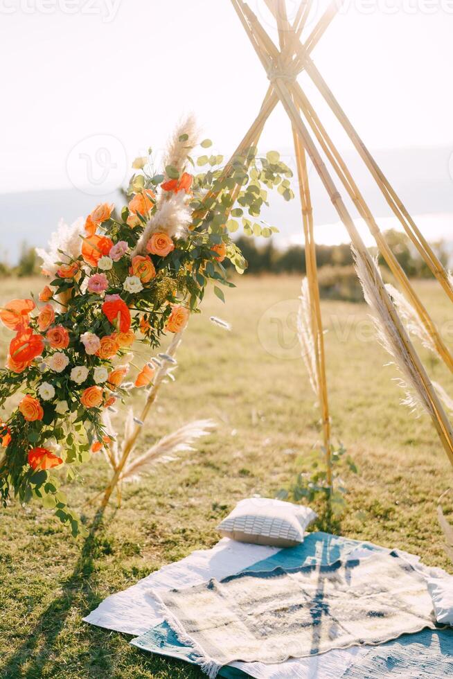 Wedding tipi arch decorated with red flowers stands on a green lawn above bedspreads and pillow photo