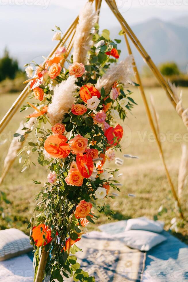 Wigwam-style wedding arch decorated with red flowers stands on a green lawn photo