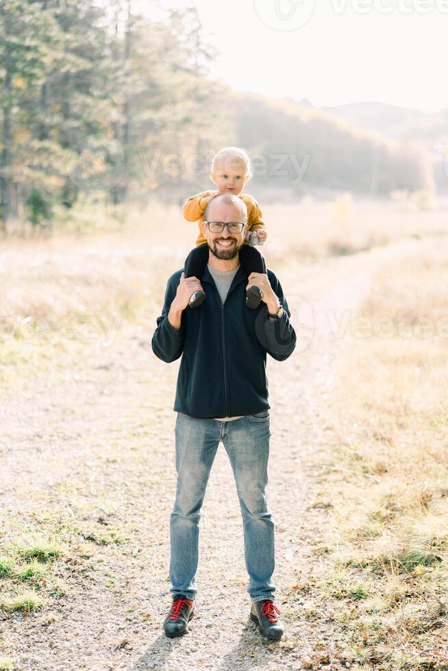 Smiling dad with a little son on his shoulders stands in a sunny forest photo