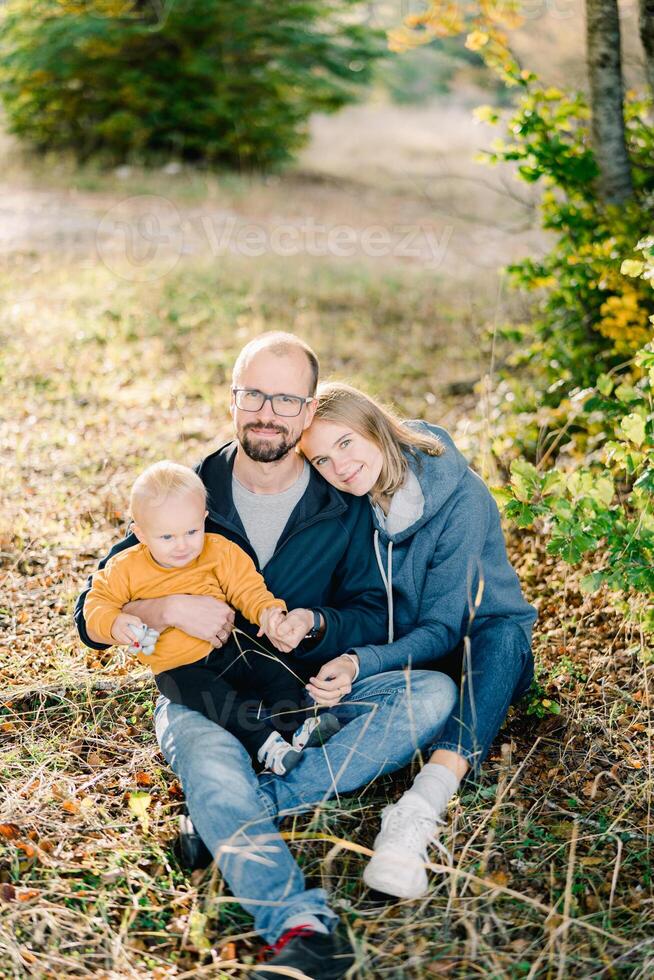 Mom rested her head on the shoulder of dad sitting with his little son on his knees on dry grass photo