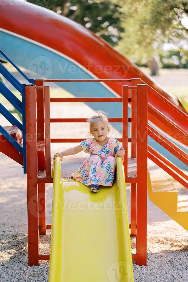 Little girl sits on top of a slide on the playground holding on to the railings photo