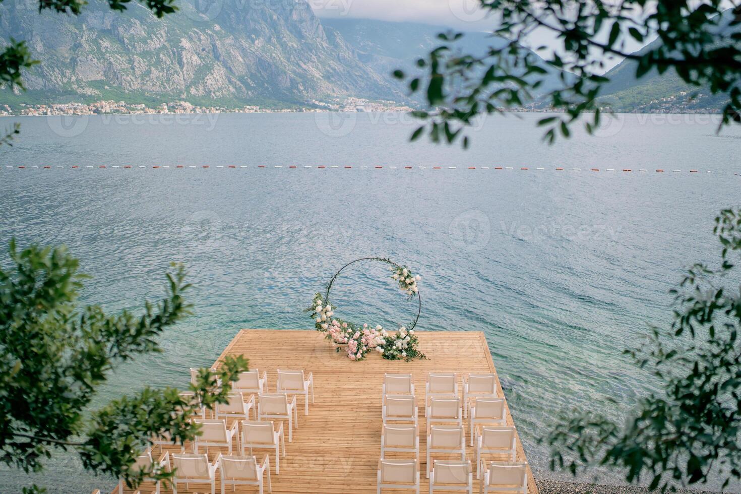 Round wedding arch stands on a wooden pier in front of rows of white chairs photo