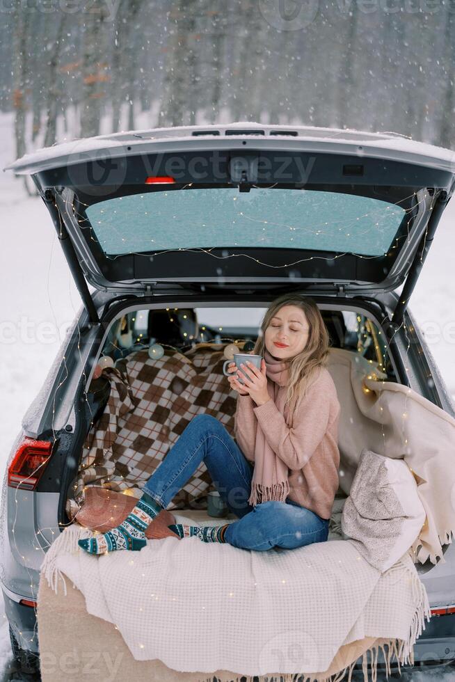 Young woman sits with a mug of coffee squinting in pleasure in the trunk of a car in a winter forest photo