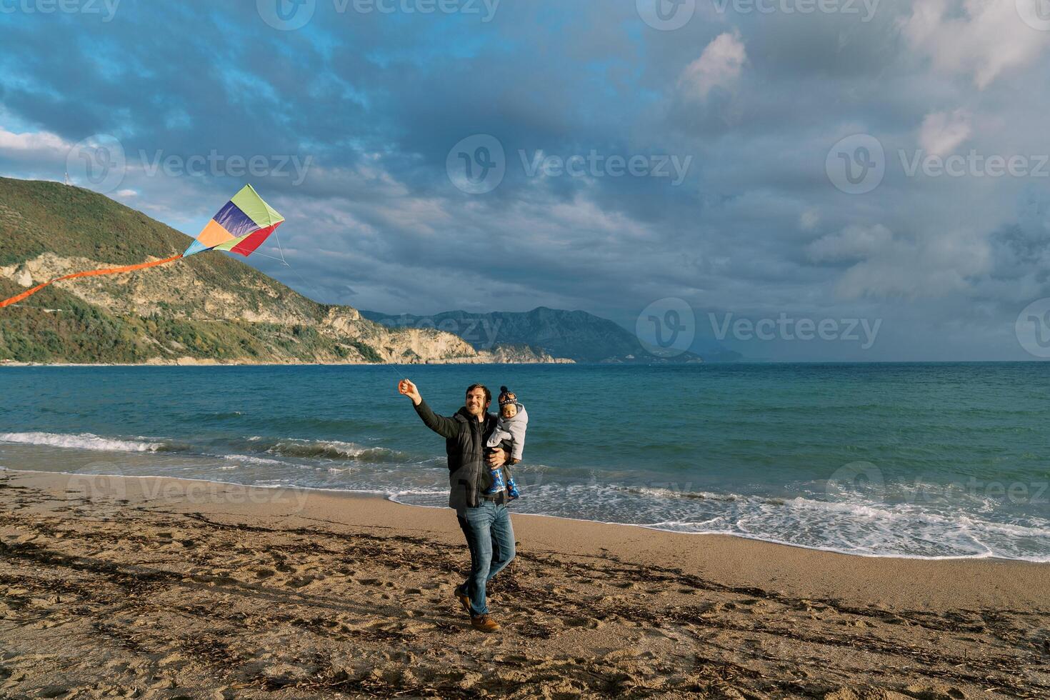Dad with a little girl in his arms looks at a hovering kite, walking with it along the seashore photo