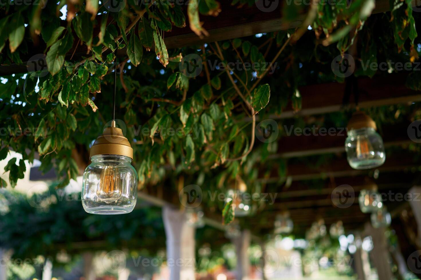 Lamps in glass jars hang from pergola beams in a green garden photo