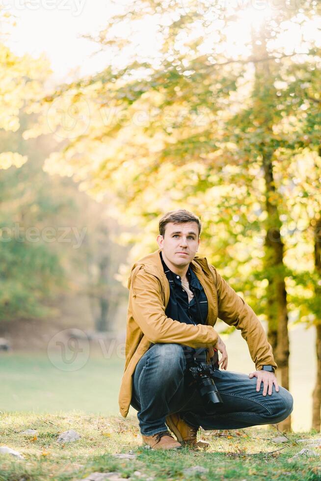 Young man with a camera around his neck squatted down in the autumn forest photo
