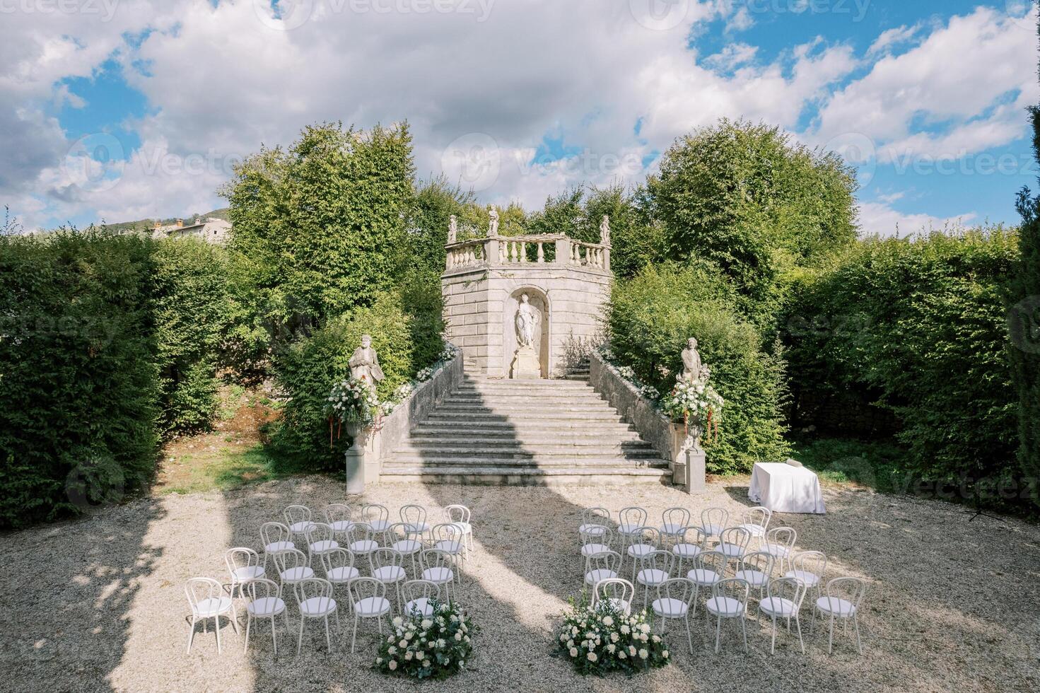 Rows of white chairs and bouquets of flowers stand in front of the steps to a nymph statue in the garden photo