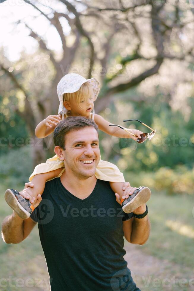 Little girl holds her dad sunglasses in her hand while sitting on his shoulders photo