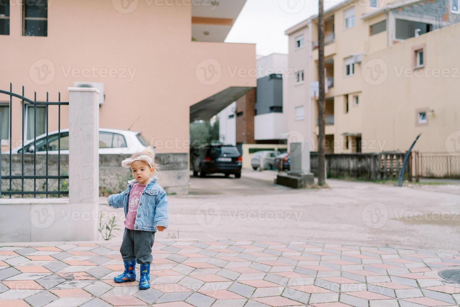 Little girl in a hoop with cat ears stands near the fence in the yard photo