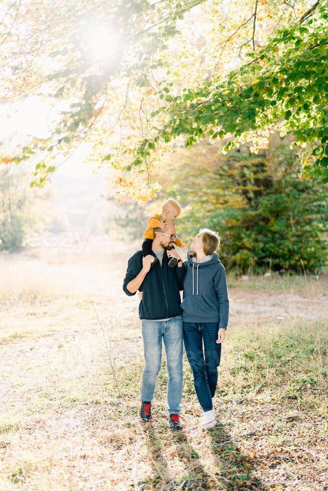 Mom hugs dad with a little boy on his shoulders, walking in the autumn park photo