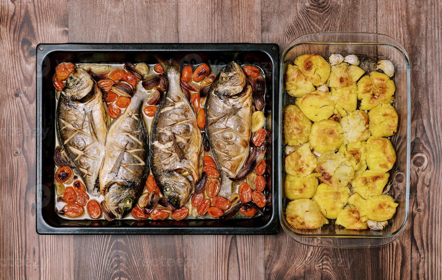 Baked sea bream with vegetables on a baking sheet stands with baked potatoes in a dish on a wooden table. Top view photo
