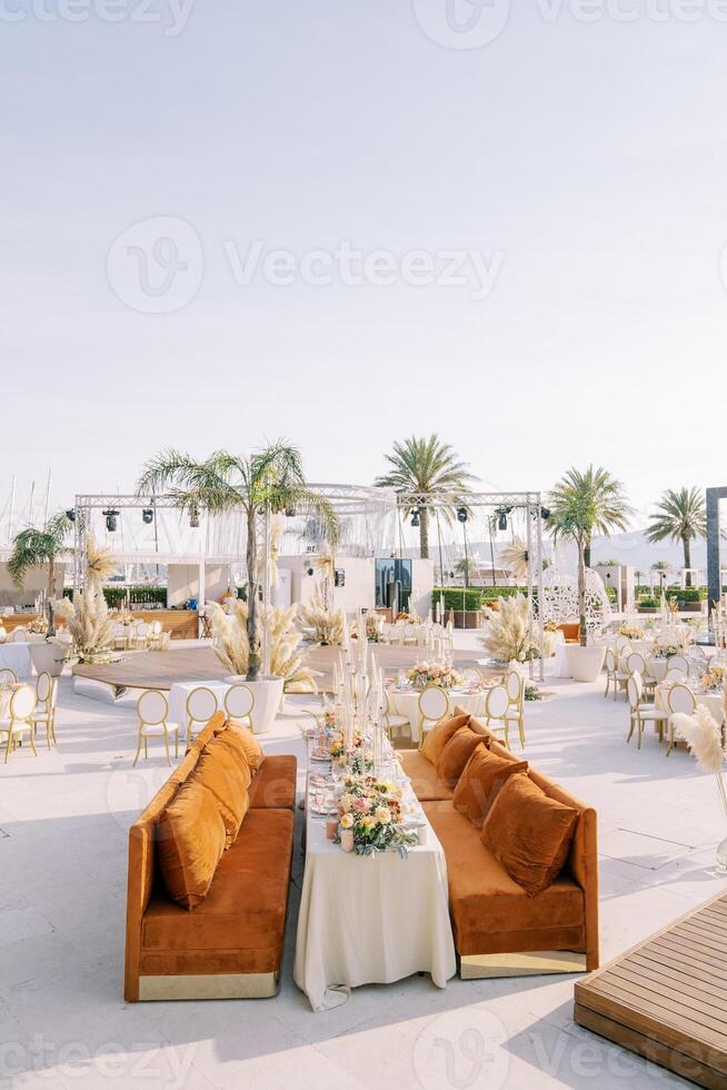 Sofas with cushions stand along a long covered table surrounded by round tables on the seashore photo