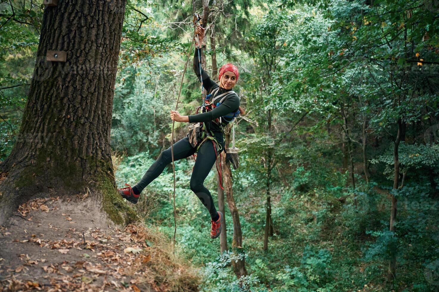 Looking, to the side, hanging on the rope. Woman is doing climbing in the forest by the use of safety equipment photo
