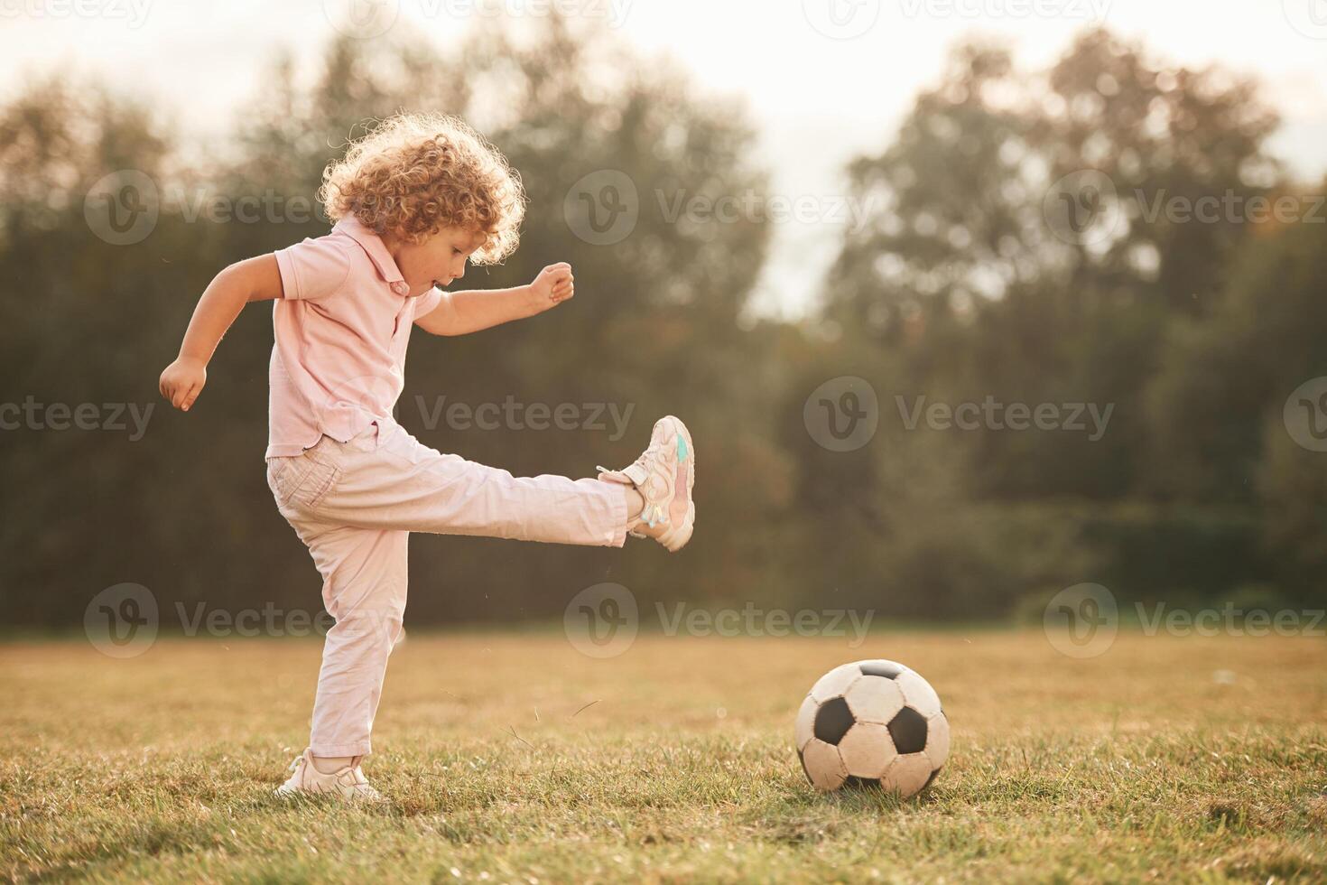 Kicks socce,r ball. Cute little girl with curly hair is playing on the field at daytime photo