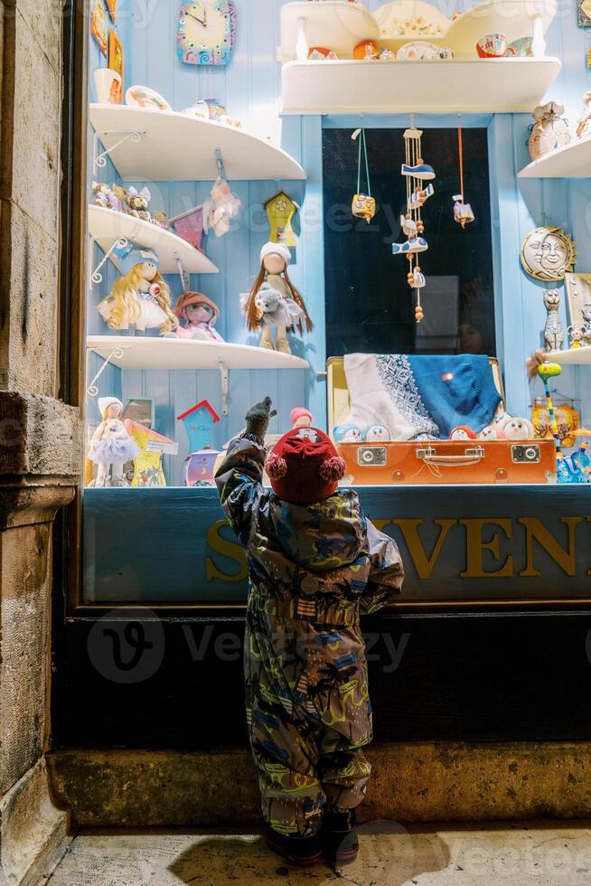 Little girl stands near a glass showcase with souvenirs and points her finger at the dolls. Back view photo