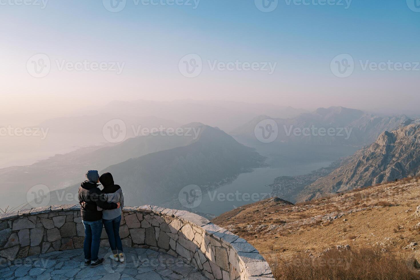 Couple of travelers in jackets hug on an observation deck over the Bay of Kotor in the fog. Montenegro. Back view photo