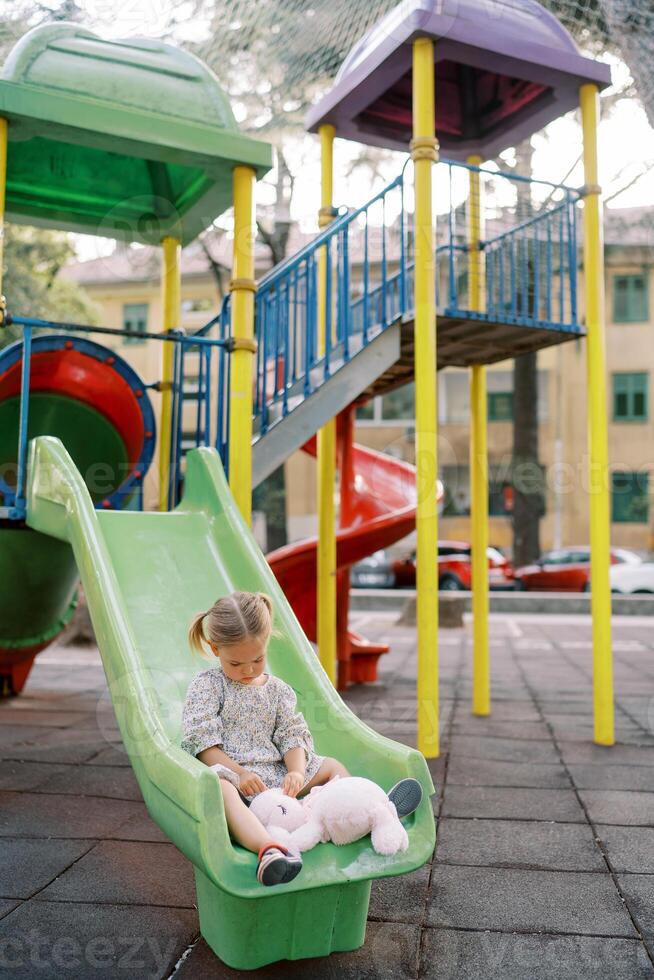 Little girl sits on a colorful slide at the playground and plays with a plush bunny photo