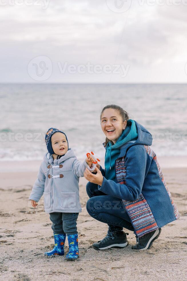 Smiling mother squat down next to a little girl with a string to a kite in her hand on the seashore photo