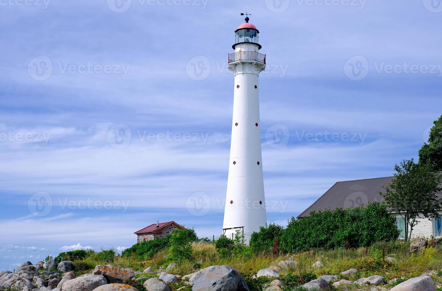 Tall white lighthouse with a red roof standing on a rocky coastal landscape under a blue sky with wispy clouds. photo