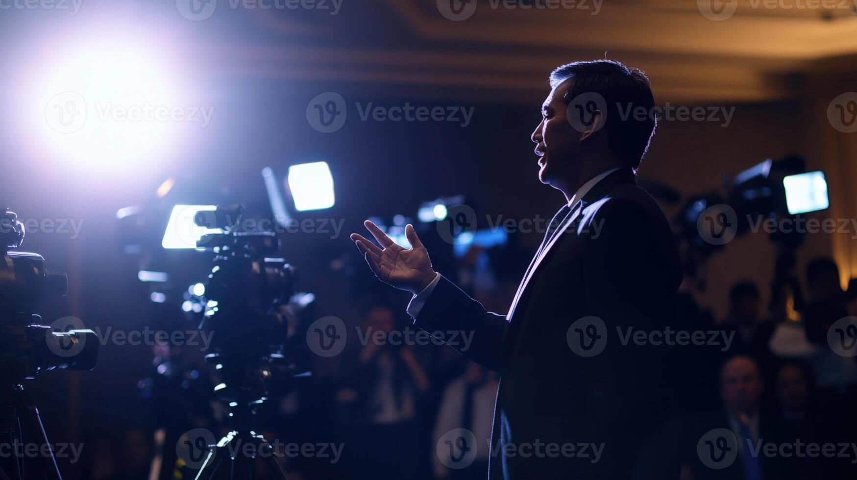 A male delegate stands confidently at the forefront gesturing passionately while addressing a sea of media personnel. The lighting casts dynamic shadows and dozens of cameras earnestl photo