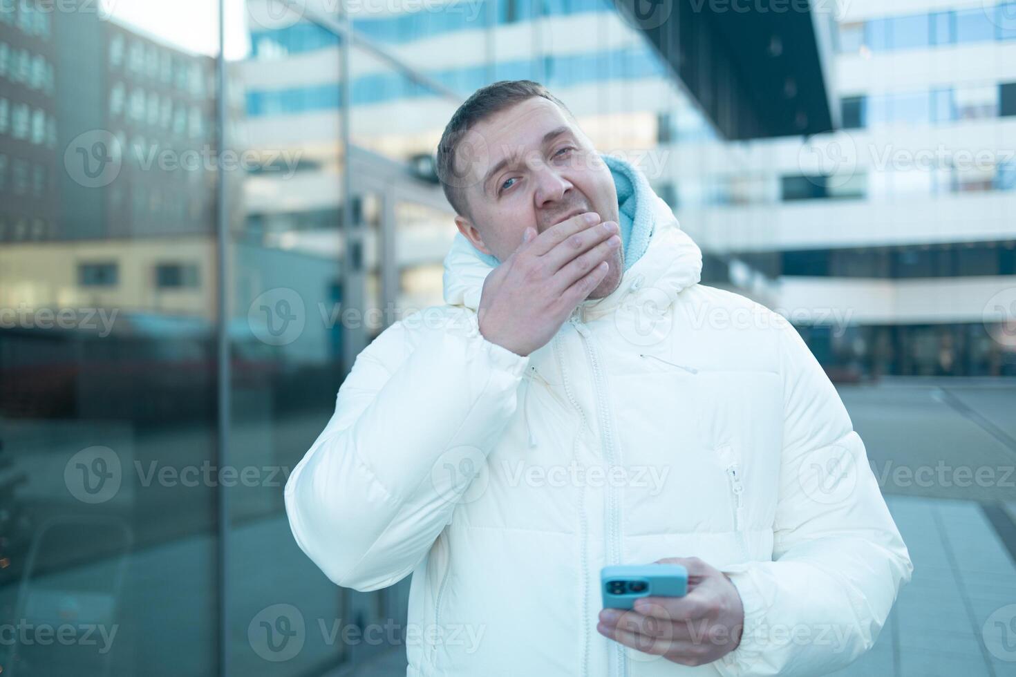Caucasian 30s man using smartphone outdoors in winter bored yawning tired covering mouth with hand. restless and sleepiness. photo