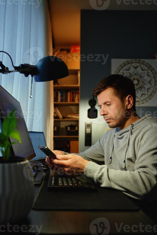 Tired freelancer procrastinating at home office, scrolling phone instead of working. Unproductive man using smartphone at home desk. Concept of procrastination and distraction in remote work photo