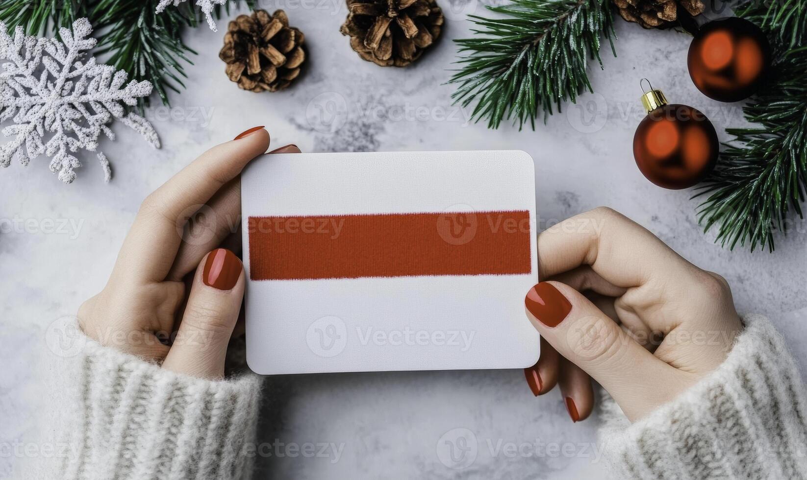 Elegant hands holding a blank gift card with red ribbon on a festive Christmas background featuring pine cones, holiday decorations and greenery photo