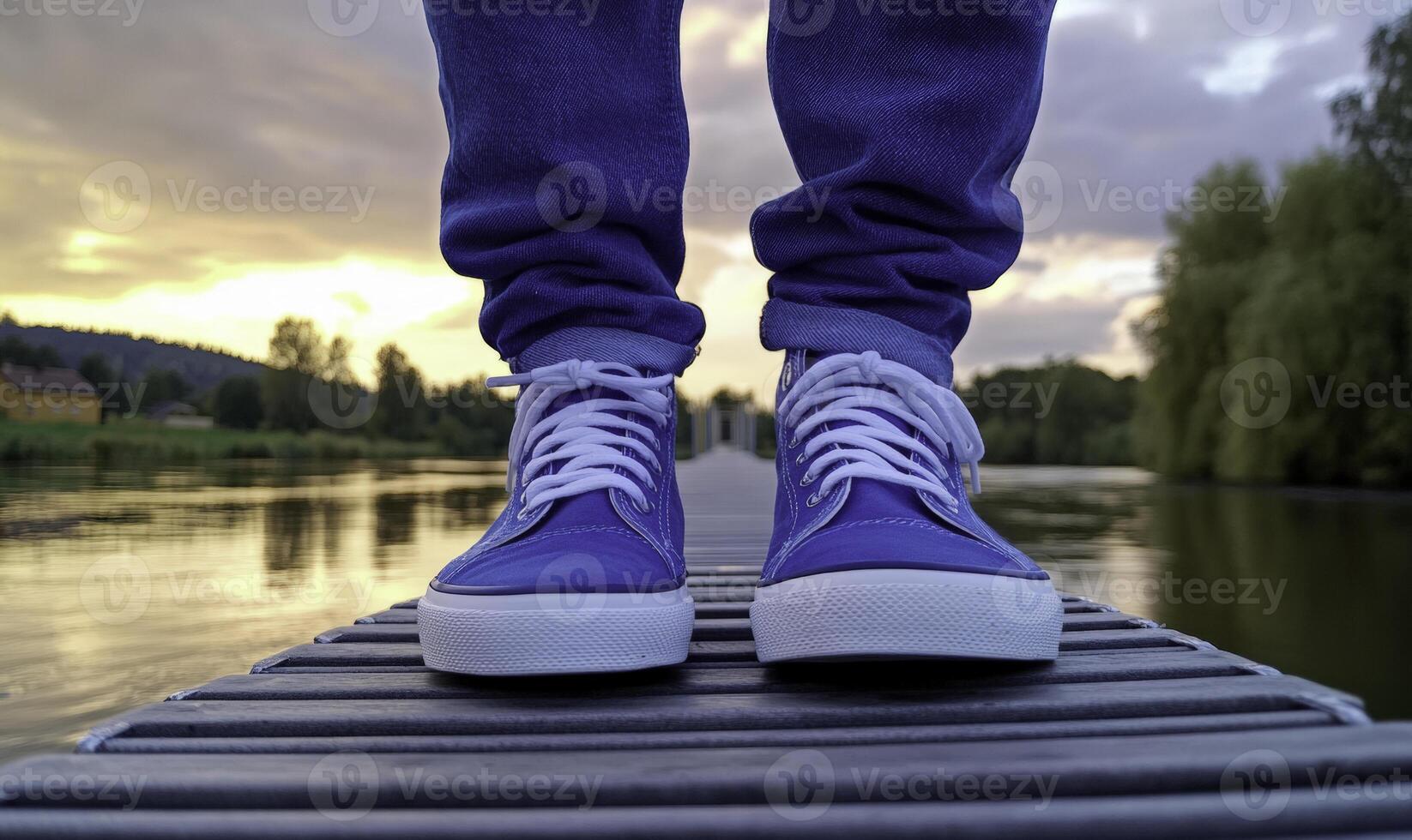 Person Standing on Dock with Blue Sneakers at Sunset Near Calm Water Surrounded by Green Trees and Dramatic Skies Reflecting Vibrant Colors photo