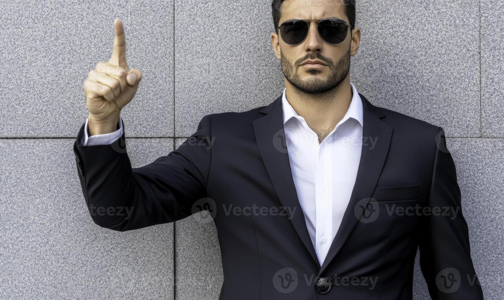 Confident Businessman in Sunglasses Gesturing with Finger Raised, Standing Against Modern Gray Wall in Professional Attire, Exuding Authority and Focus photo