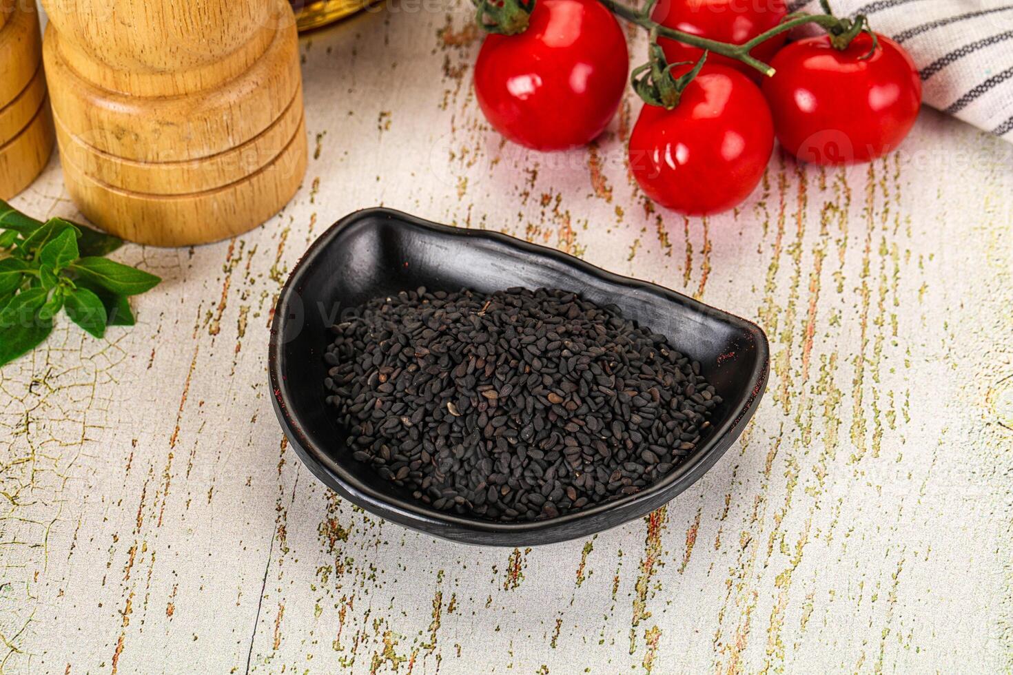 A bowl of black seeds on a table next to a knife and a tomato photo