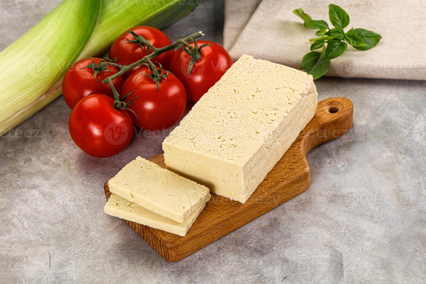 A piece of cheese on a cutting board next to tomatoes and a green leaf photo
