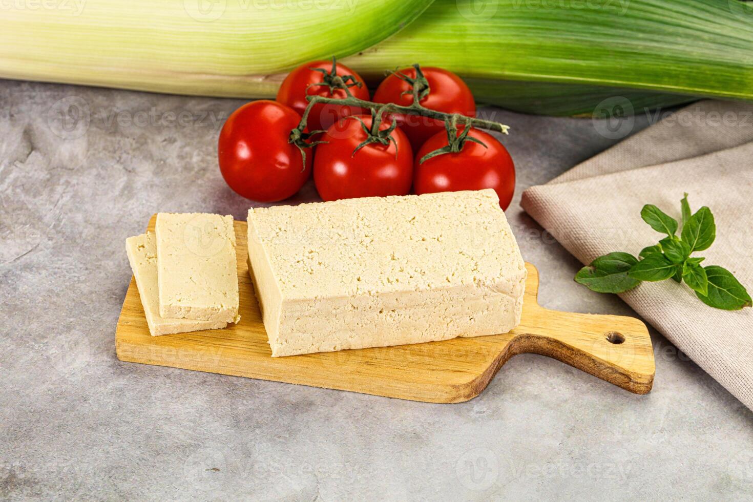 A piece of tofu on a cutting board next to a tomato and a green leaf photo
