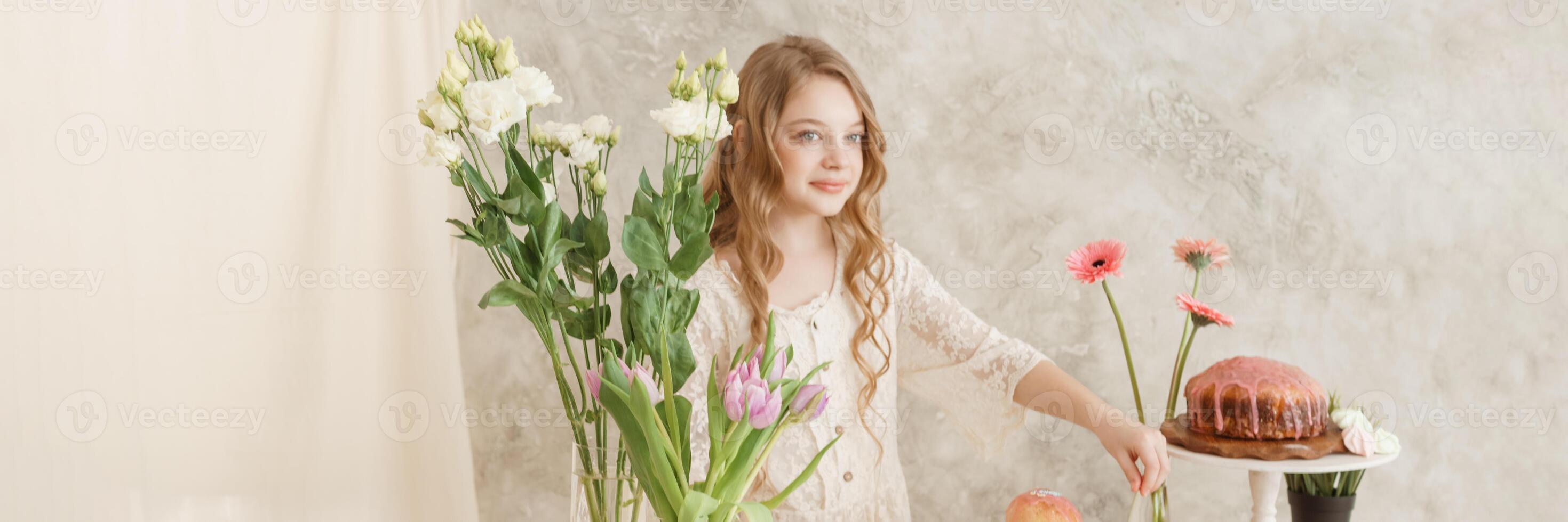 girl with long hair in a light dress is sitting at the Easter table with cakes, spring flowers and quail eggs. Happy Easter celebration. photo