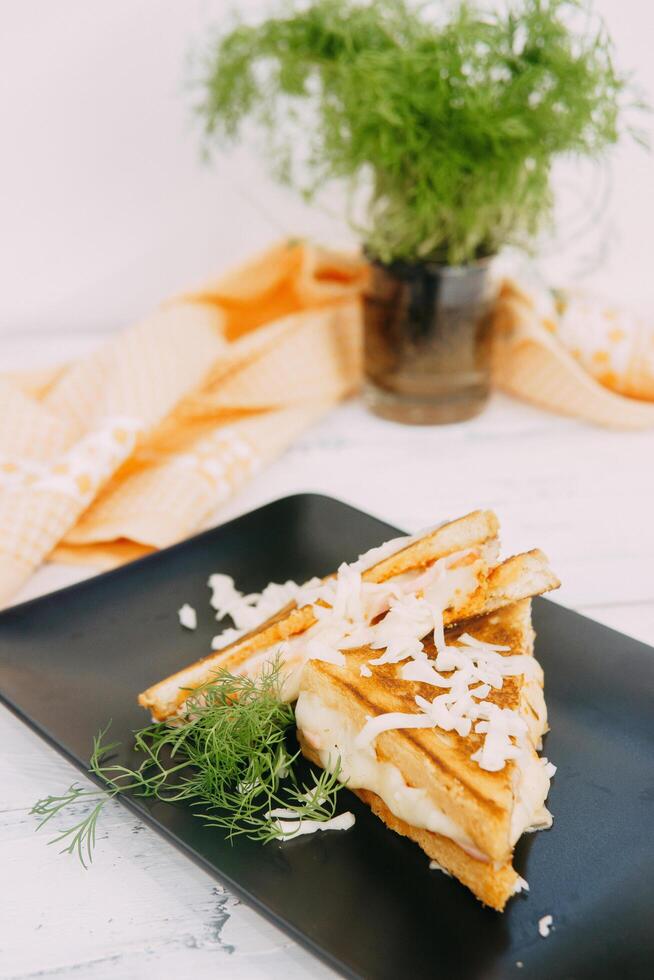 Grilled sandwich with melted cheese, served on a black plate, garnished with fresh herbs and shredded cheese, set against a cozy kitchen background featuring yellow cloth and herbs. photo