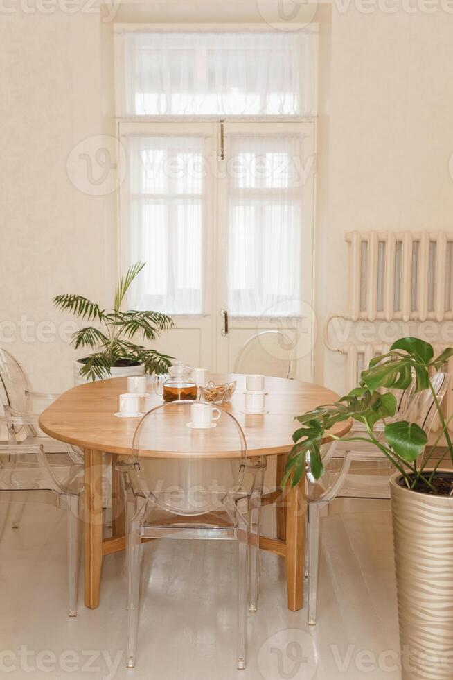 Cozy dining setting featuring round wooden table surrounded by transparent chairs, complemented by lush green potted plants, creating a warm and inviting interior atmosphere. photo