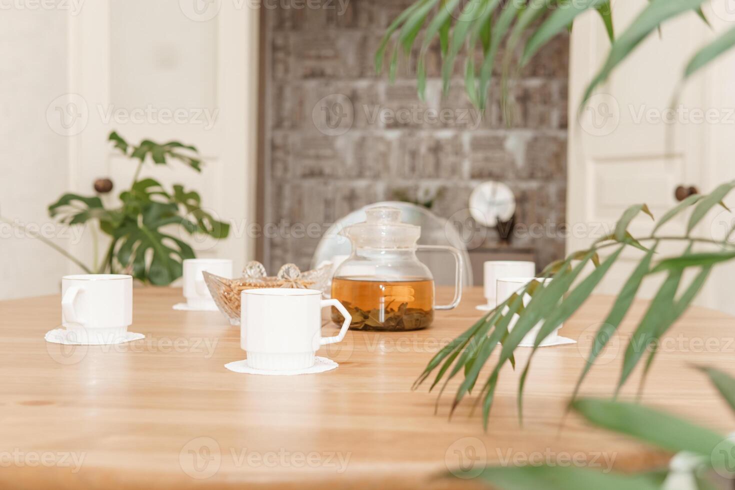 cozy composition featuring a teapot and cups on a round wooden table, complemented by green plants and soft lighting in tastefully designed interior space. photo