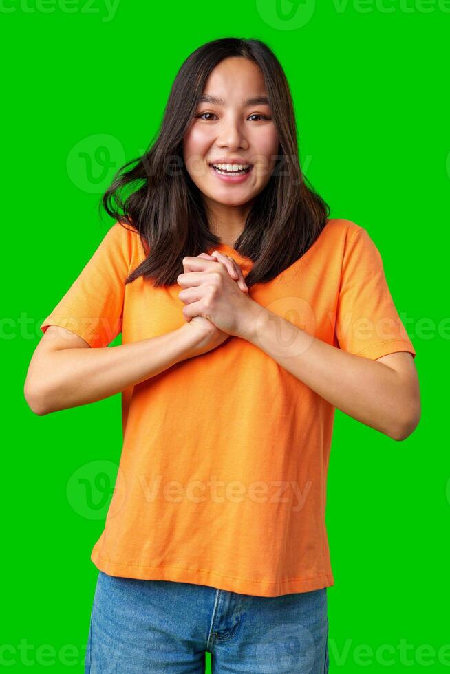 Young woman expresses excitement with joyful smile and hands together, wearing an orange shirt against a bright background photo