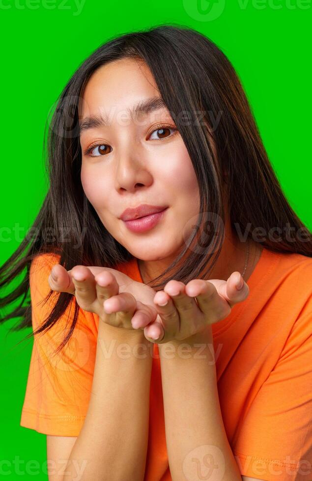 Young woman in orange shirt blowing a kiss against a bright green background while smiling warmly photo