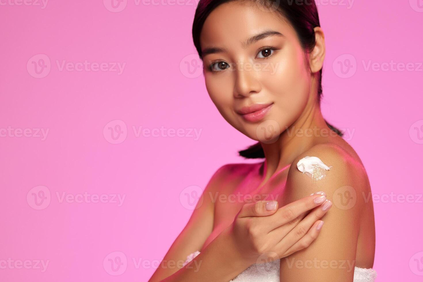 Young woman applying skin cream against a vibrant pink background during a beauty routine session photo