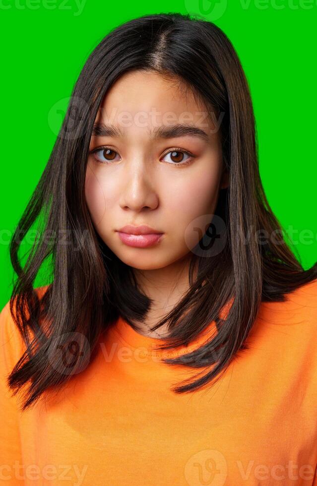 Teen girl with long dark hair looks thoughtfully ahead against a bright green background wearing an orange shirt photo