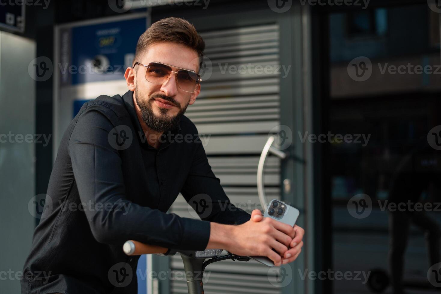 A young happy hipster man with an electric scooter standing in front of a modern business building on the city street and using a mobile phone. photo